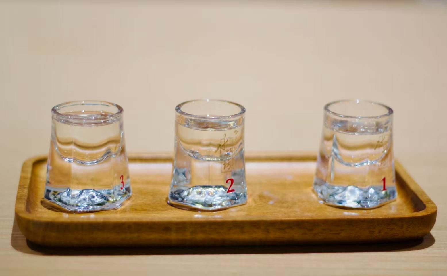 Three clear shot glasses with wide thick bottoms that taper to a narrower top sit on a wood serving tray, each filled with different aged baijiu