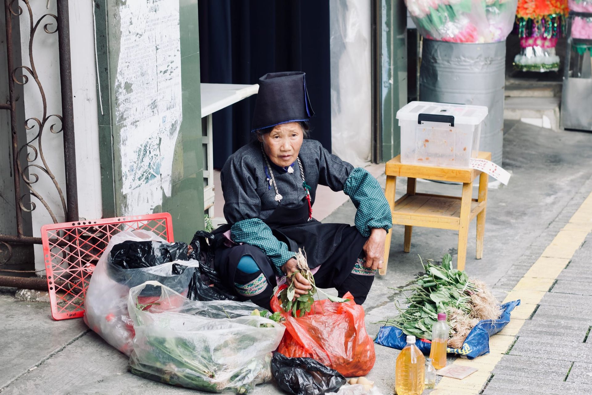 A Miao woman in traditional charcoal dress and silver jewelry sits on the roadside selling her produce from plastic bags.