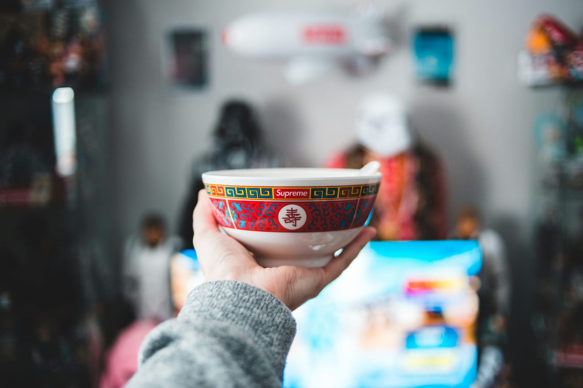 A hand holding a steaming Supreme bowl with Chinese art deco on the side, aloft.