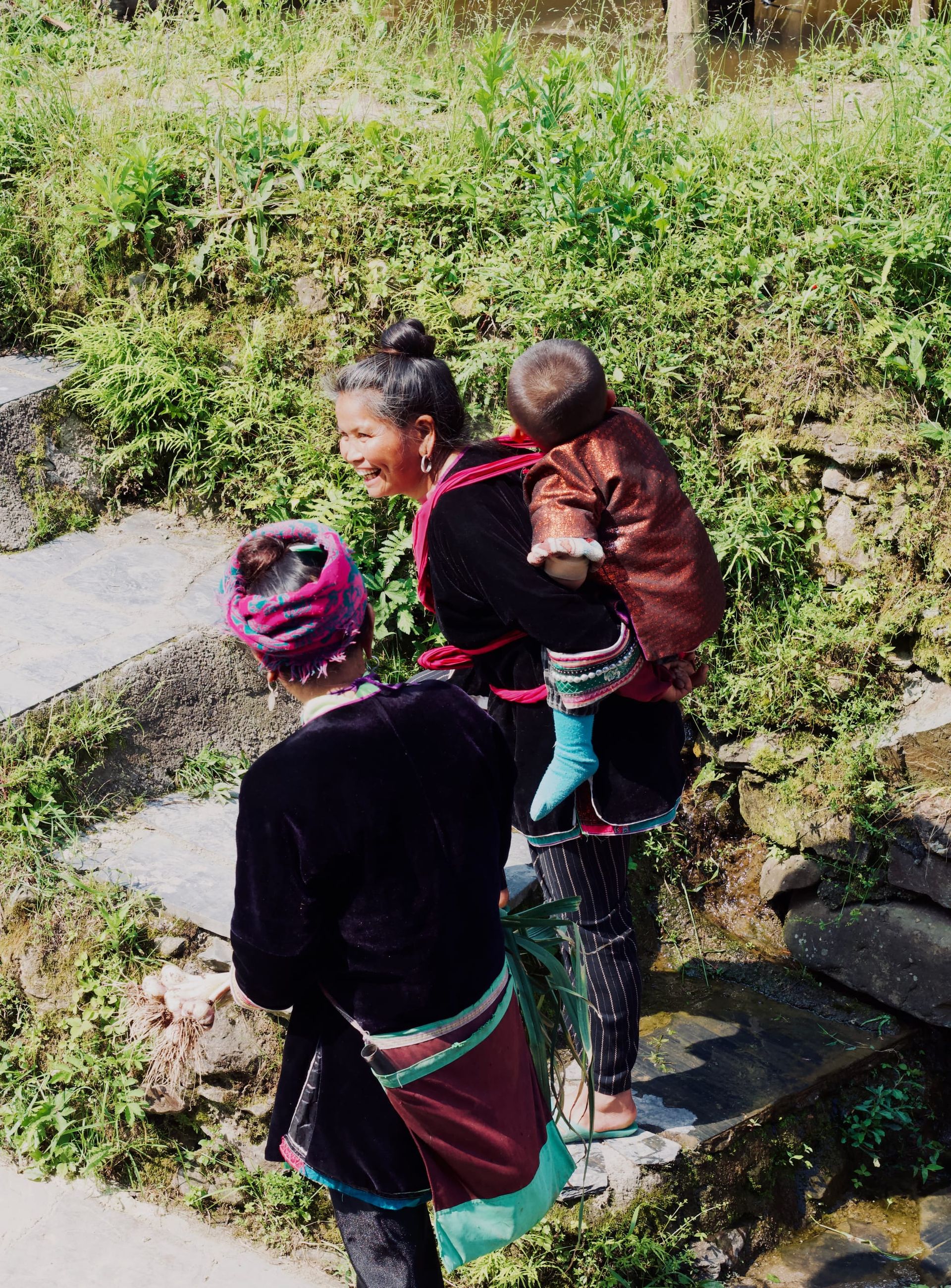 Two elder Dong women walk along a stone path, one carrying a baby Dong on her back wearing traditional attire. 