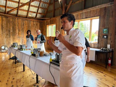 Arnold standing in front of a demonstration table setup with hot plates and pots and sets of ingredients, leading a cooking class as four volunteers stand behind waiting for instructions.