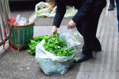A customer bends over to pick up a giant bag of coriander leaves next to other leafy greens they purchased, ready to put them into a bamboo backpack on the side of the stree