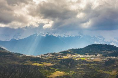 A photo of Shangri-La - a green forested plateau in front of snow-capped mountains with gray clouds above with sporadic sunrays piercing through.