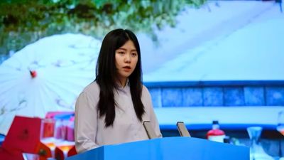 Yifei Guo speaking at a sky blue podium, with a Chinese market behind her.