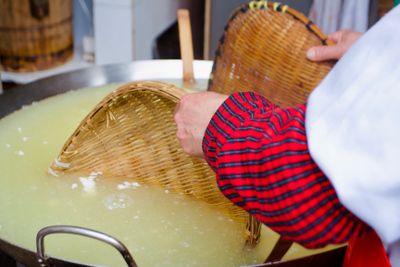 Yellow-ish clear water with small white clouds in a large stainless steel vat; a worker uses bamboo trays to wisk the water in the tofu molding process.