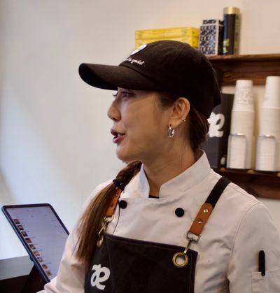 A photo of Ally Jiang, a baker and entrepeneur, standing in her chefs outfit and apron at the checkout counter of her bakery