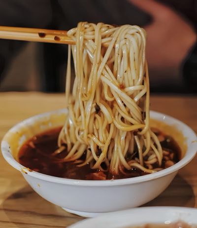 A white bowl filled with redish-brown Guaiwei sauce and noodles being held up out of the bowl by chopsticks.