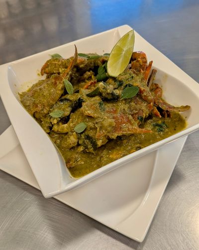 A square white bowl filled with crab curry - a green chunky dish, with a wedge of lime on the side.