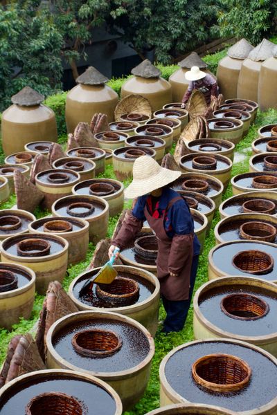 A Chinese worker tends to soy sauce fermentation casks by scooping out the existing soy sauce and pouring it back over the beans it was extracted from, amongst many rows of open casks.