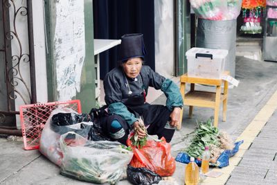 A Miao woman in traditional charcoal dress and silver jewelry sits on the roadside selling her produce from plastic bags.