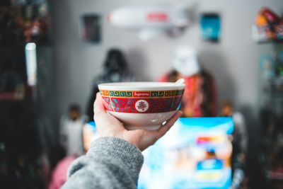 A hand holding a steaming Supreme bowl with Chinese art deco on the side, aloft.