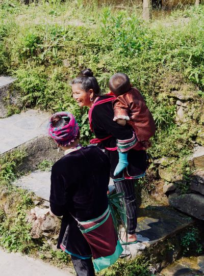 Two elder Dong women walk along a stone path, one carrying a baby Dong on her back wearing traditional attire. 