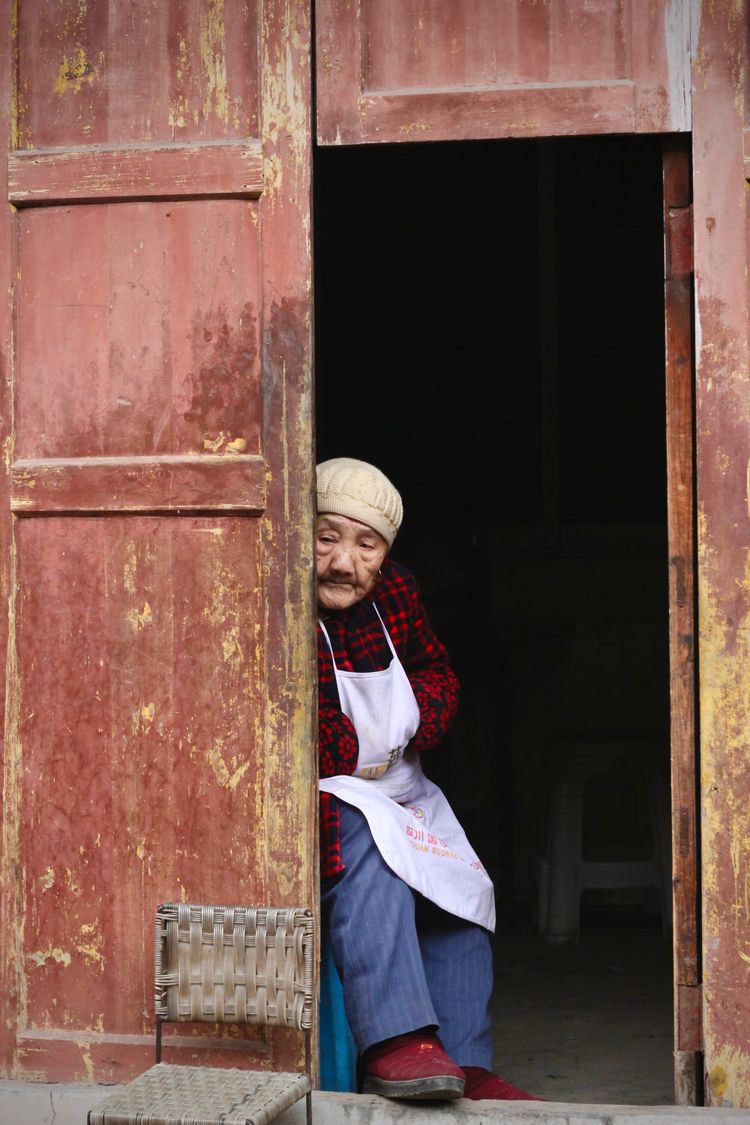 An elderly Sichuan, Chinese woman sits in the faded red wooden doorway to her shop in Yaoba Historic Village.