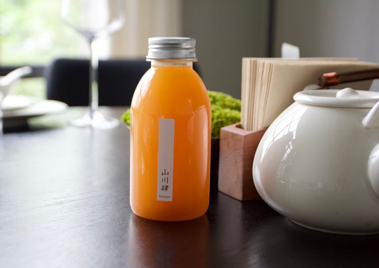 A small bottle of orange liquid sits on a dining table. A small white label with the brand Shan Cheng He in Chinese characters is on the side of the bottle.