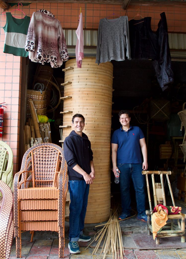 Two European white males stand next to a 10-foot large stack of giant bamboo steamer baskets in a storage area.