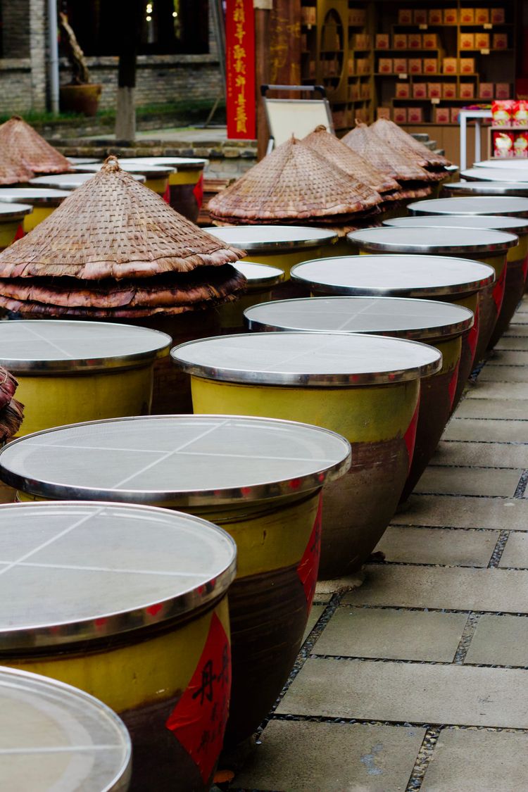 Rows of soy sauce urns sit on a tiled patio, their lids replaced with a white mesh to keep out insects while the contents aerate. 