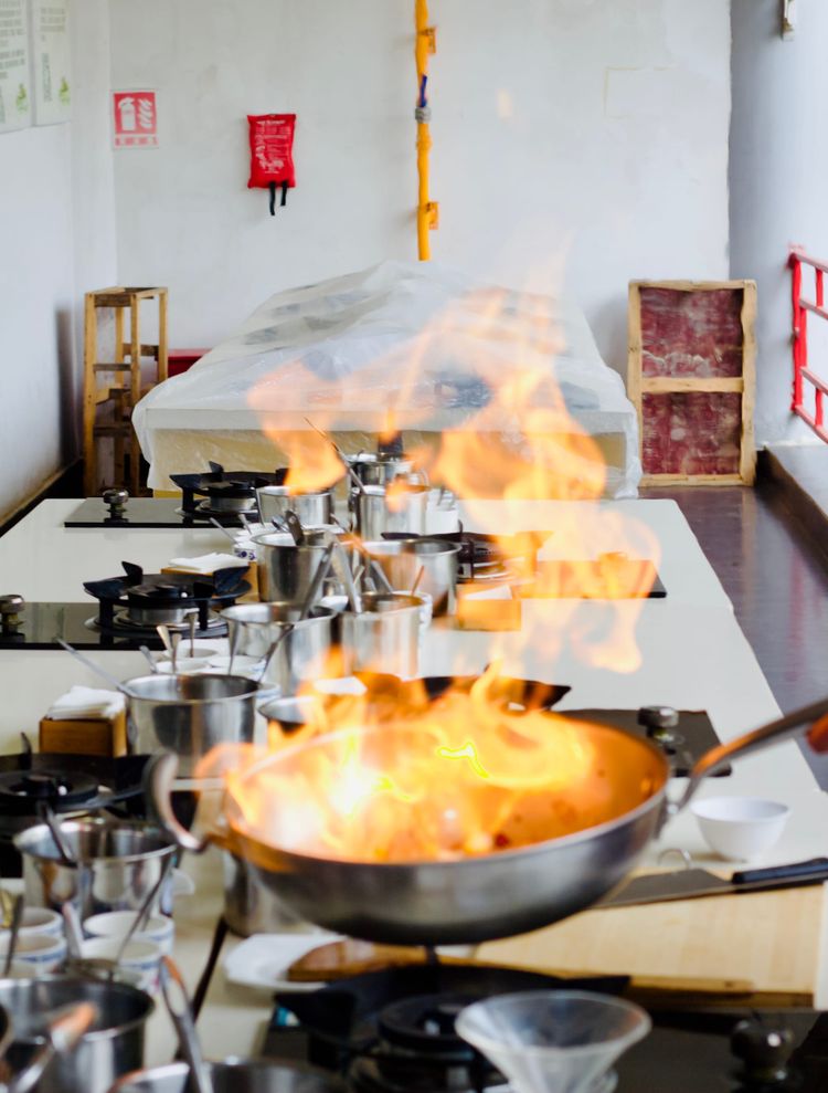 A stainless steel wok is alit with bright orange fire as the chef flambés the dish in a professional kitchen.