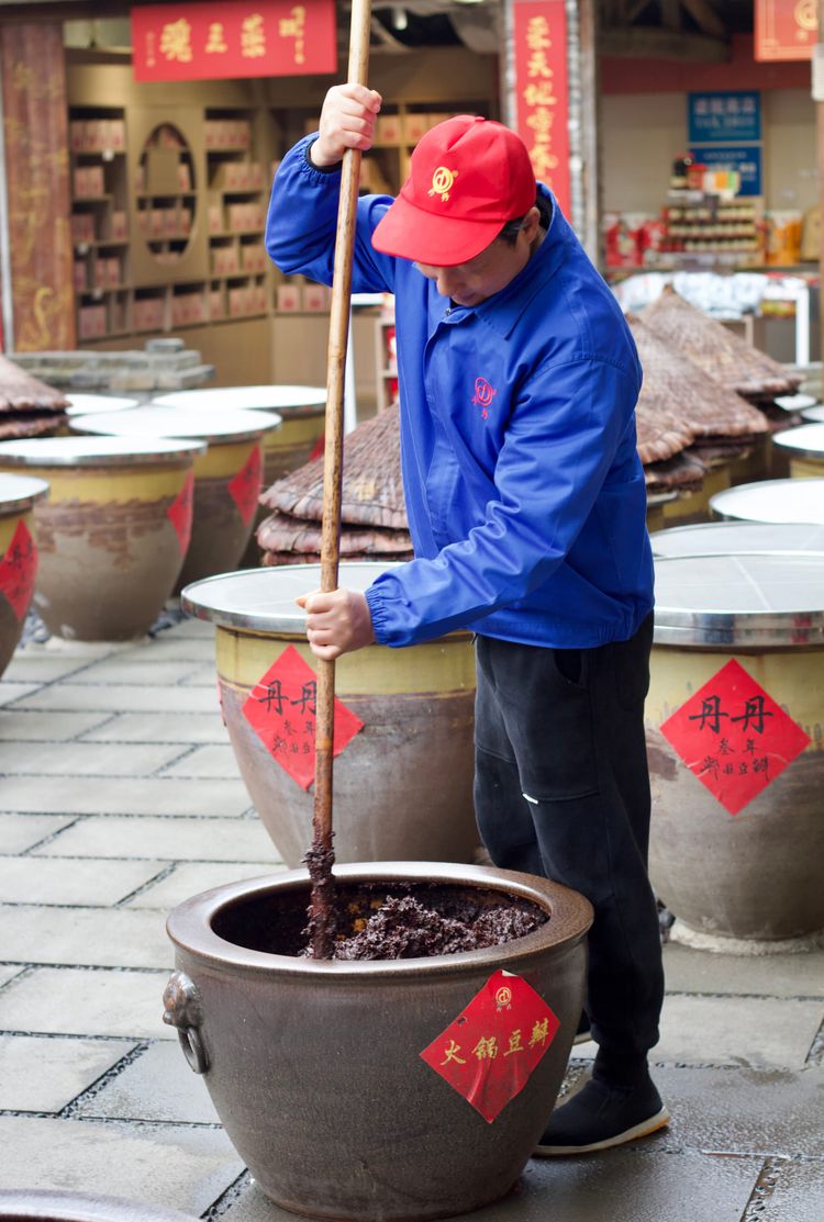 A shifu uses a bamboo stick to stir a large ceramic urn filled with doubanjiang on a stone patio, next to other urns fermenting.