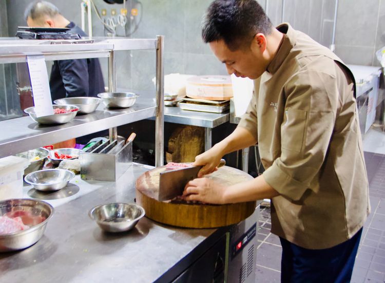 Chef Tang, using a meat cleaver on a large circular wood cutting board inside a typical stainless steal professional kitchen.