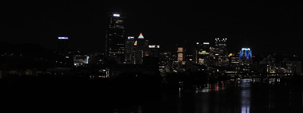 Pittsburgh at night. Skyscrapers and city lights glow in the night and reflect off the river, shrouded by trees, buildings, and the 16th street bridge.