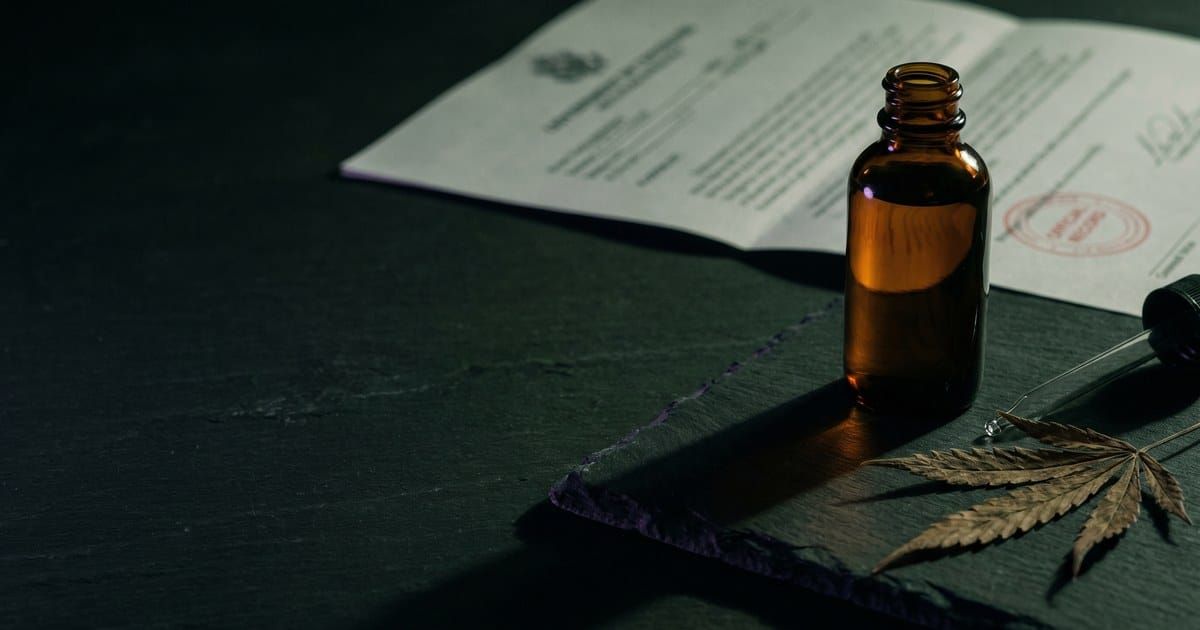 An amber CBD tincture bottle and a dried hemp leaf on a dark surface, with a stamped government document blurred behind it.