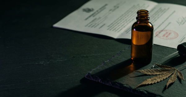 An amber CBD tincture bottle and a dried hemp leaf on a dark surface, with a stamped government document blurred behind it.