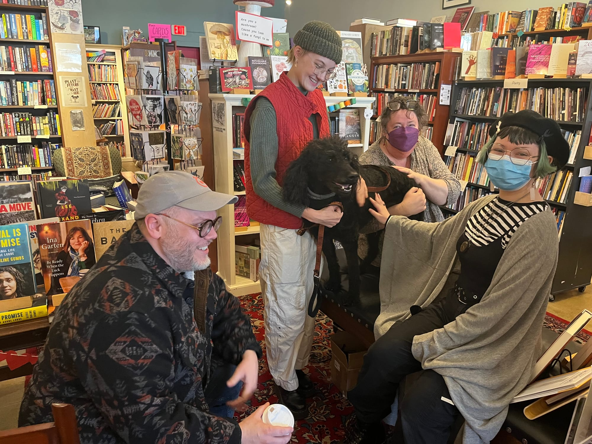 Four people gathered around an adorable black dog in a bookstore. Kelly is wearing a purple KN95 and Arianna is wearing a blue surgical mask.