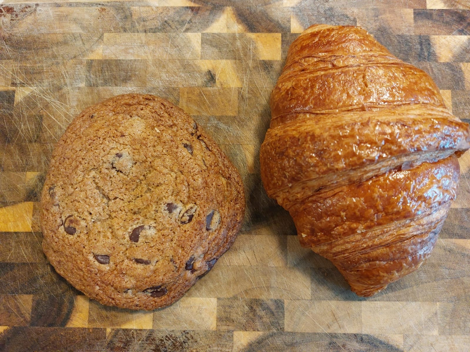 Chocolate chip cookie and croissant, both relatively large and deep in color, lying side by side on a cutting board