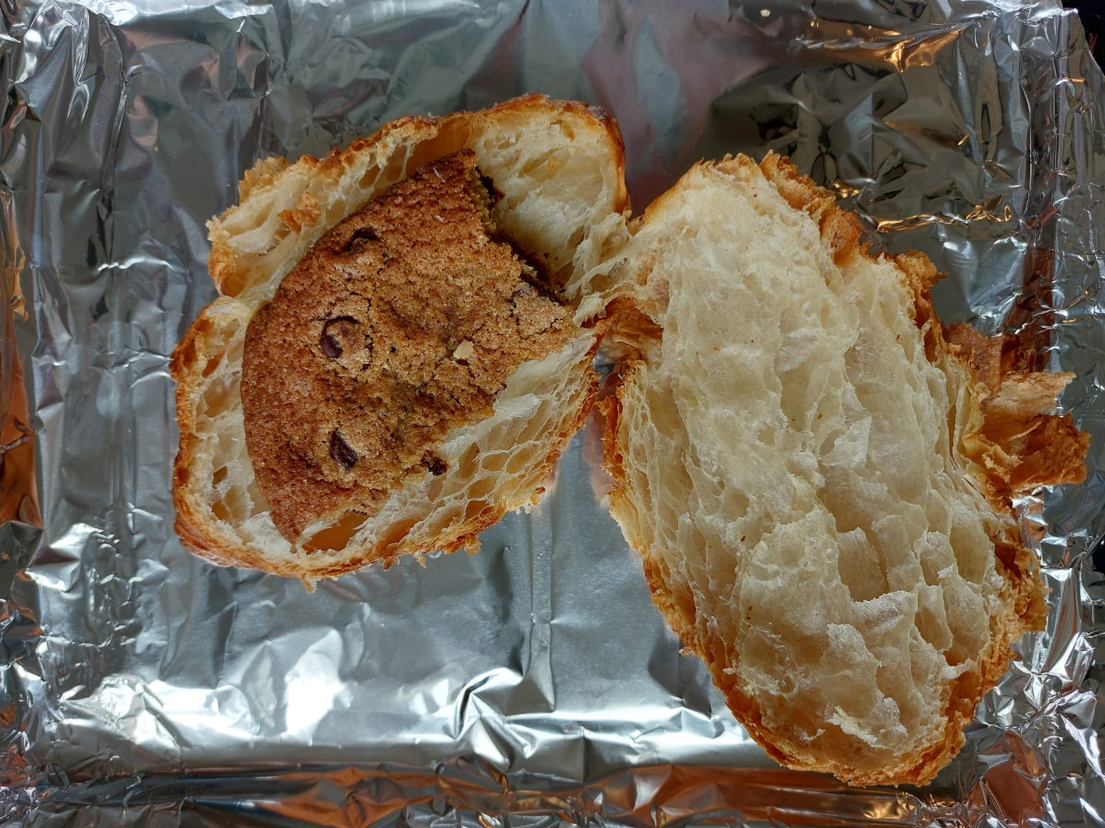 Croissant lying cut open on a baking sheet lined with aluminum foil, a segment of cookie placed on top of one side