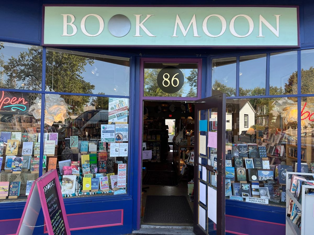 Front entrance of Book Moon Books, with whimsical exterior color scheme of pale green, dark blue, pink, and purple, books displayed in windows, and door swung open.