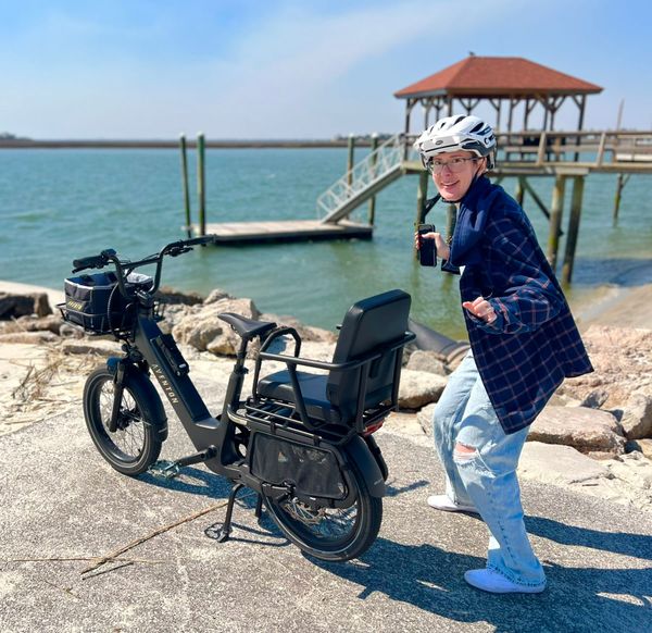 Patricia Lockwood in a bicycle helmet standing next to a bike with a seat on the back, near a body of water