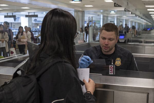 A U.S. border agent wearing a dark uniform sits behind a metal counter at an airport. He is wearing a light blue glove as he prepares to accept a document from a traveler with long, black hair who is facing him across the counter.