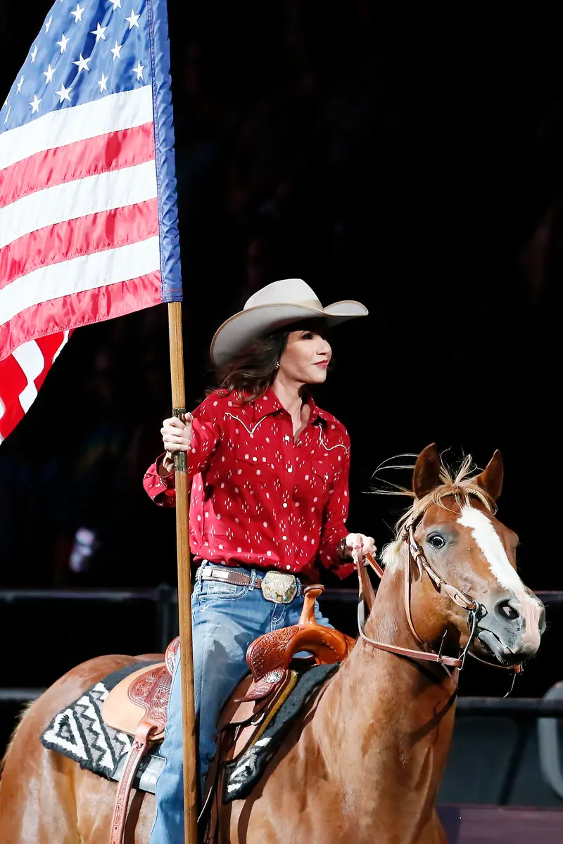 Kristi Noem, in a cowboy hat, Western shirt and jeans, holds an American flag while riding a horse.