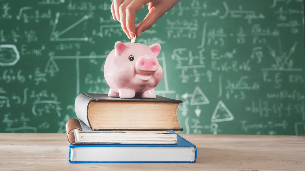 A stack of books is sitting on a table. A pink piggybank is on top of the stack of books. A hand is putting coins into the piggybank.