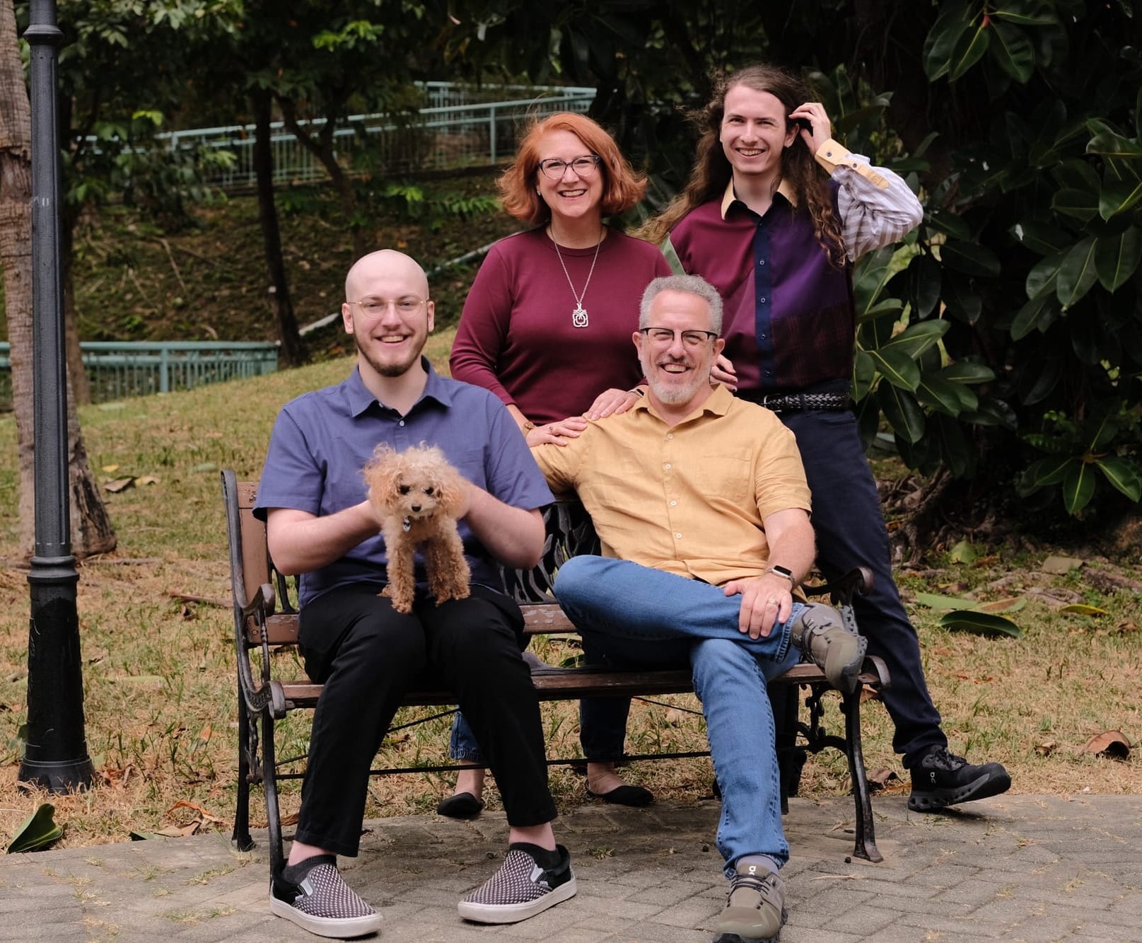 Two men sit on a park bench. One holds a small poodle. Behind them stand a woman and another man. All are smiling as they look at the camera.