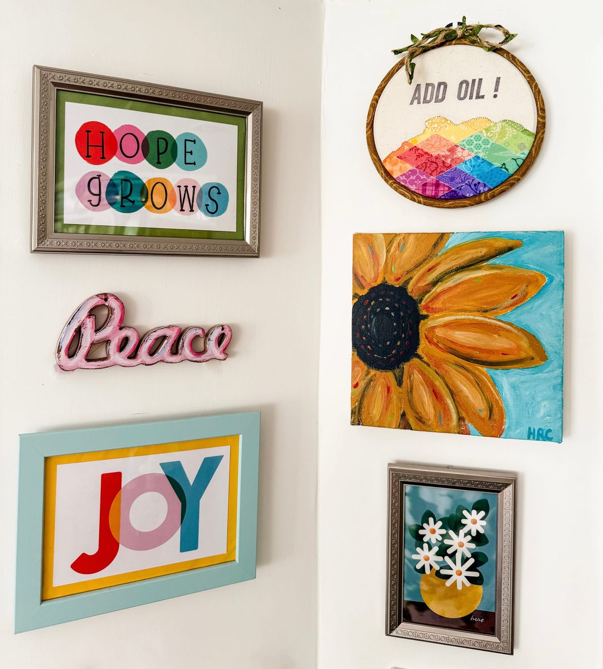 Six pieces of colorful art on a white wall. Text on the art: Hope Grows, Add Oil, Peace, Joy, and Here. 