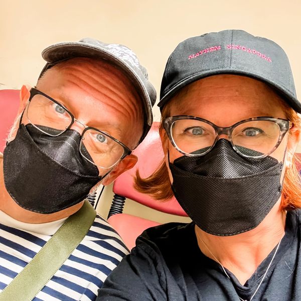 A man and woman wearing glasses sit in a hospital waiting room with masks on. 