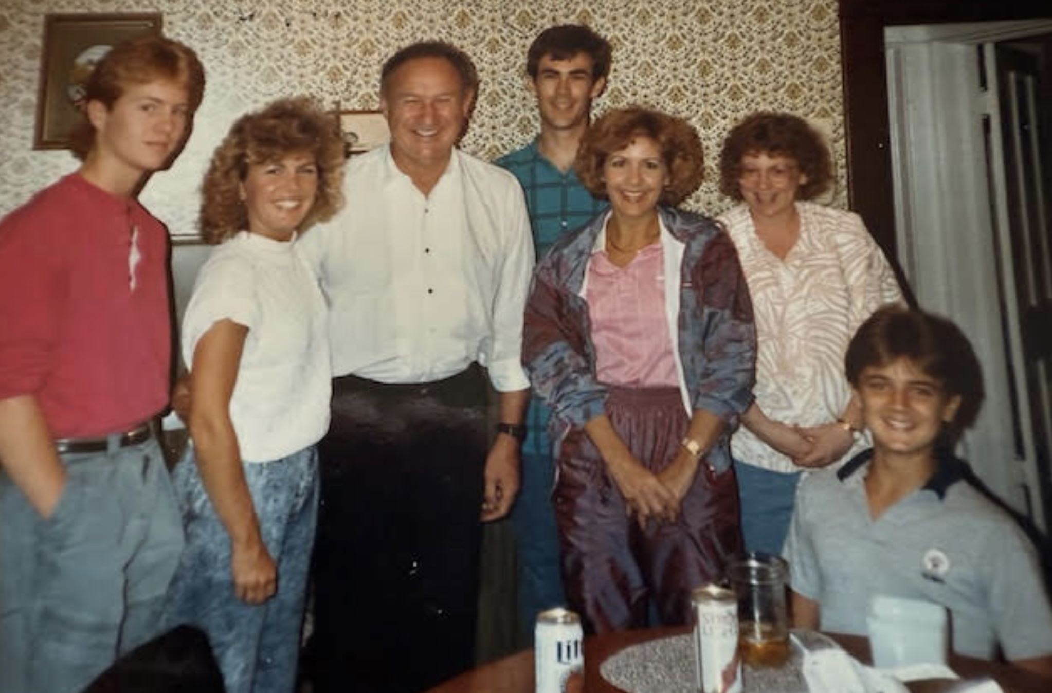 Gene Hackman with a group of people in a 1980s kitchen. Beers on the table. Hackman is wearing a tuxedo, jacket off, tie off, shirt unbuttoned