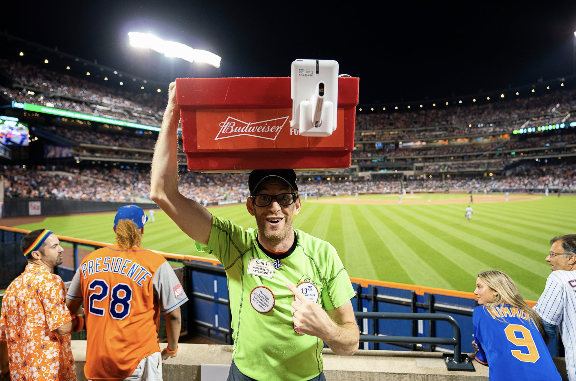 A beer vendor at a Mets game, looking at the camera, giant beer cooler balancing on his head