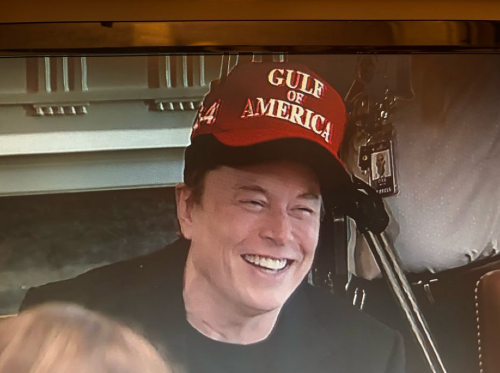 Elon Musk wearing a read "Gulf of America" cap on top of his usual black "Make America Great Again" cap. He's smiling like he's made the best joke ever.