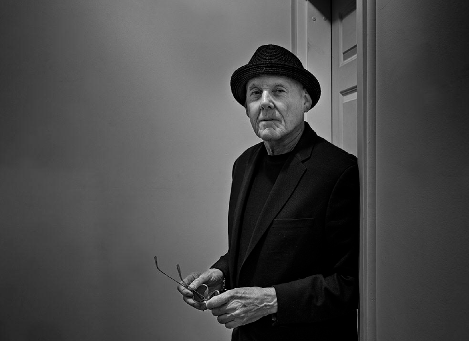 Photo of the author, Billy Price. Man in a black suit jacket, black shirt, and a black hat, holding sunglasses, leaning against a door and looking at the camera