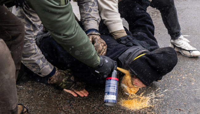 A person being held on the pavement by three ICE agents. They are completely helpless as one of the agents unloads a can of pepper spray into their eyes at point blank range. 