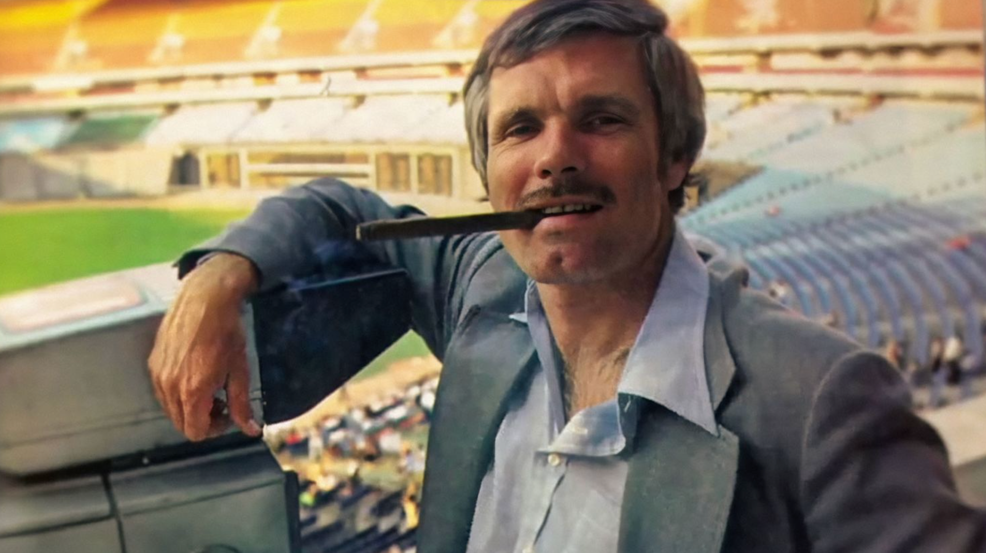 A youngish Ted Turner leaning on a camera in Fulton County Stadium, a cigar hanging out of his mouth