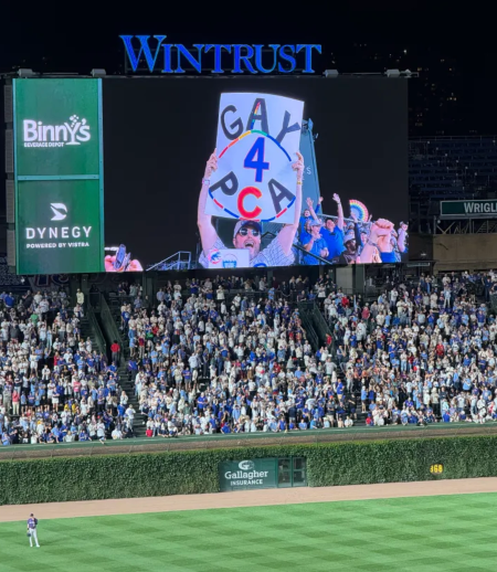 Photo of the Wrigley Field outfield, bleachers, and scoreboard. On the scoreboard is a fan holding up a sign that says "GAY 4 PCA"