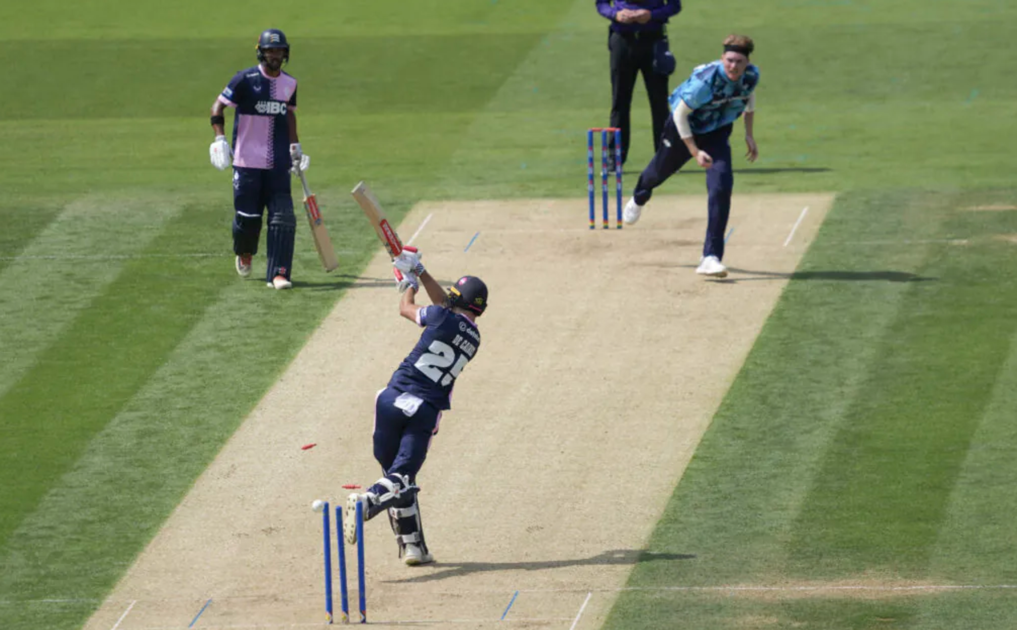 A cricket bowler gets one past a batter, knocking bails off the wickets, as the batter is at full extension