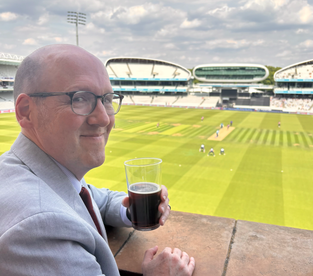 Me, in a suit and tie holding a beer, while standing on the terrace of the Lord's Cricket Ground Pavilion, with cricket action going on in the background
