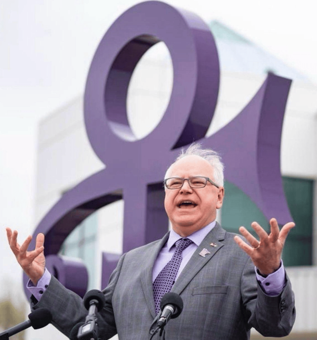 Tim Walz in front of Paisley Park studios, and a giant purple Prince symbol, wearing a suit with a purple shirt and a purple tie 