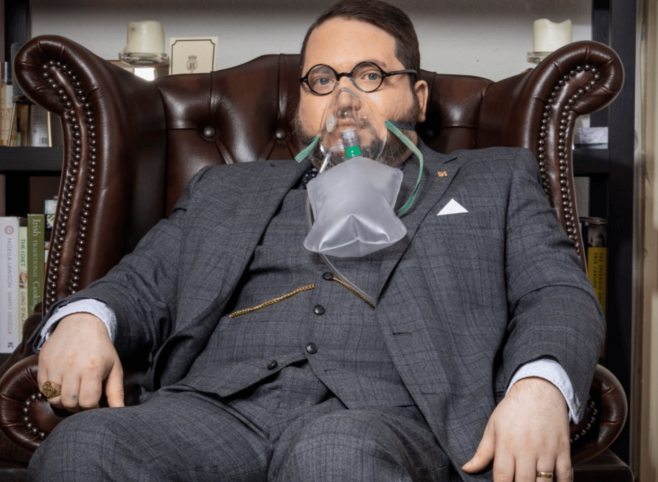Nicholas Rossi pretendig to be Arthur Knight, sitting in a large leather armchair, wearng a posh British suit, and wearing an oxygen mask
