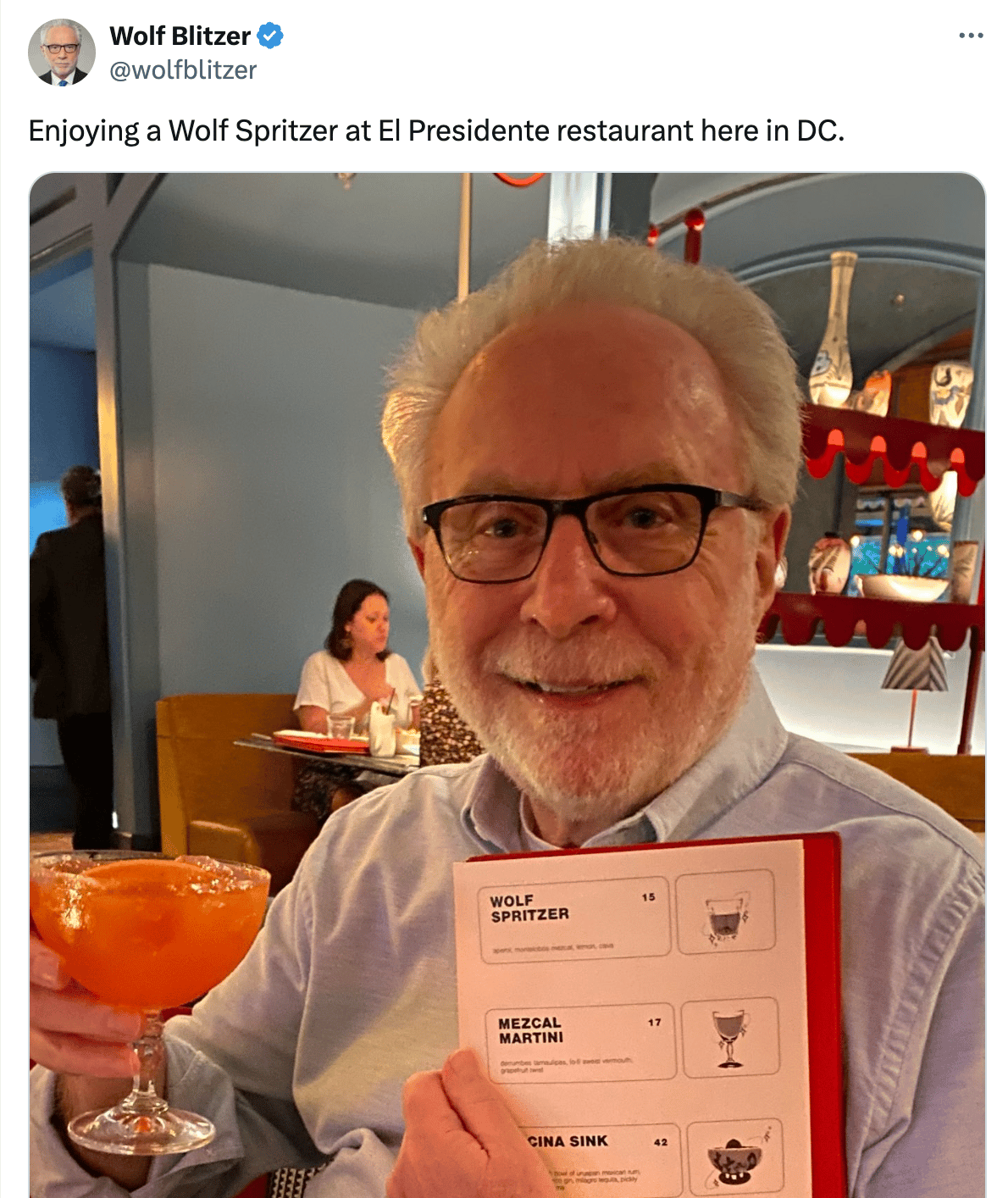 Tweet from Wolf Blitzer: "Enjoying a Wolf Spritzer at El Presidente restaurant here in DC." he's holding up the drink and a menu showing that it is indeed called a Wolf Spritzer