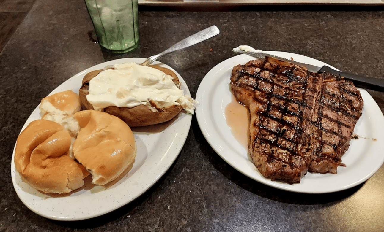 A t-bone steak on one plate, a baked potato and a roll on the other
